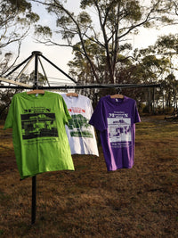 Three t-shirts in green, white, and purple hanging on a clothesline outdoors with trees in the background.