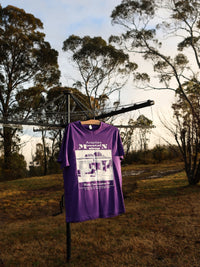 Purple t-shirt with white graphic design hanging outdoors on a clothesline with trees in the background