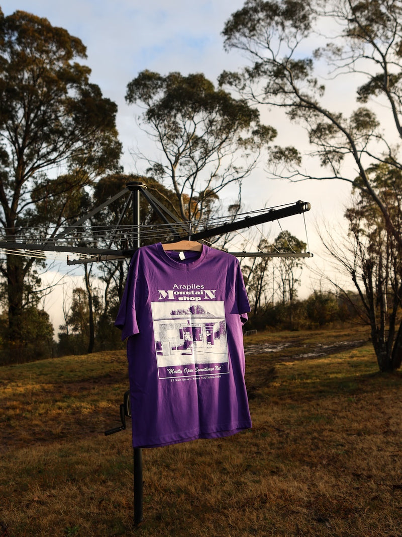 Purple t-shirt with white graphic design hanging outdoors on a clothesline with trees in the background