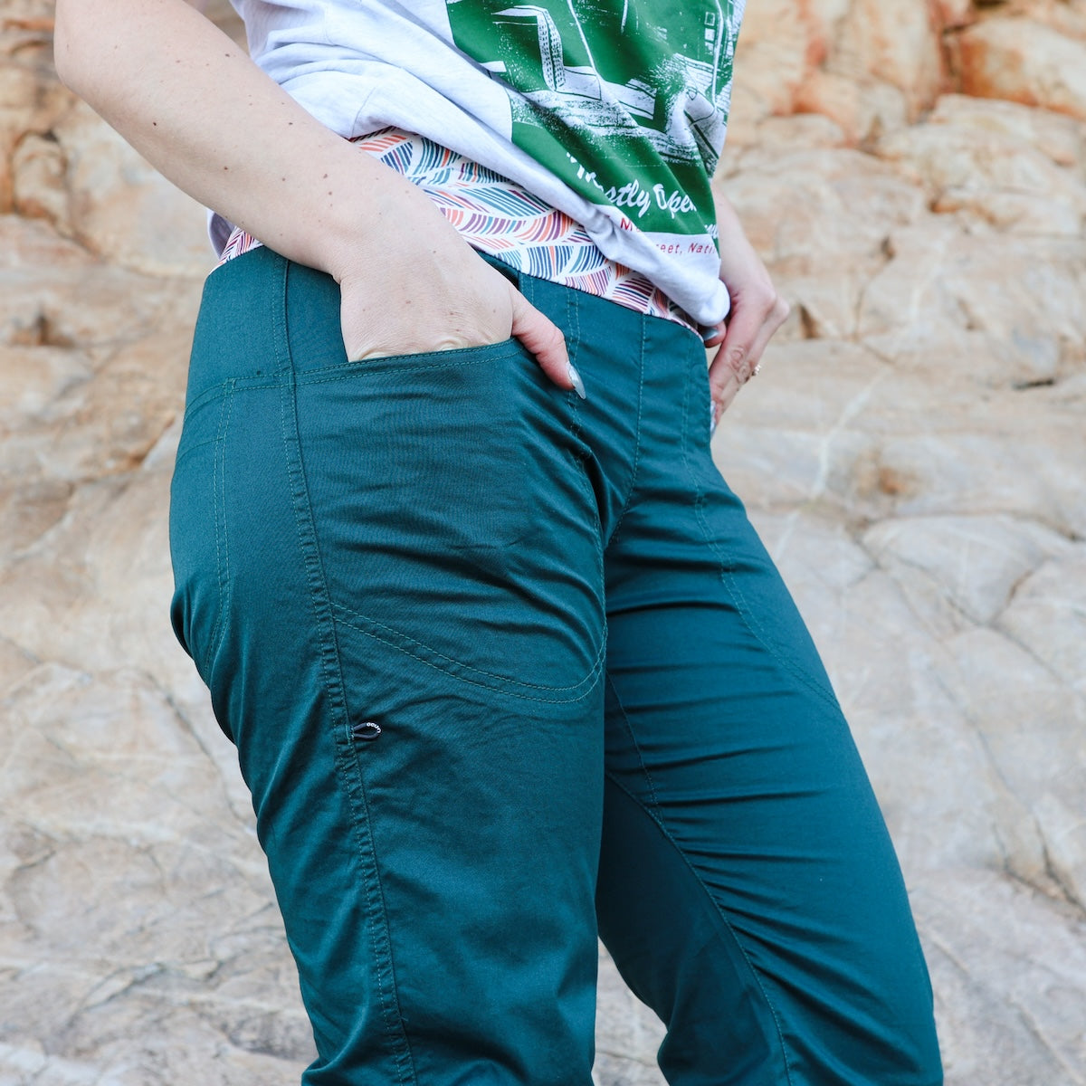Person wearing teal pants with a patterned shirt in front of a textured beige wall.